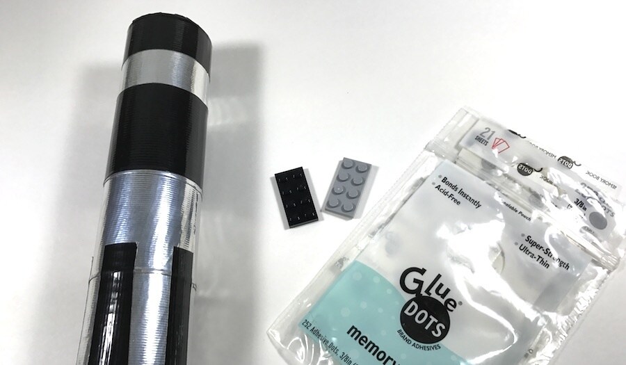 A glass bud vase partly wrapped in duct tape to resemble a lightsaber, next to a package of Glue Dots.
