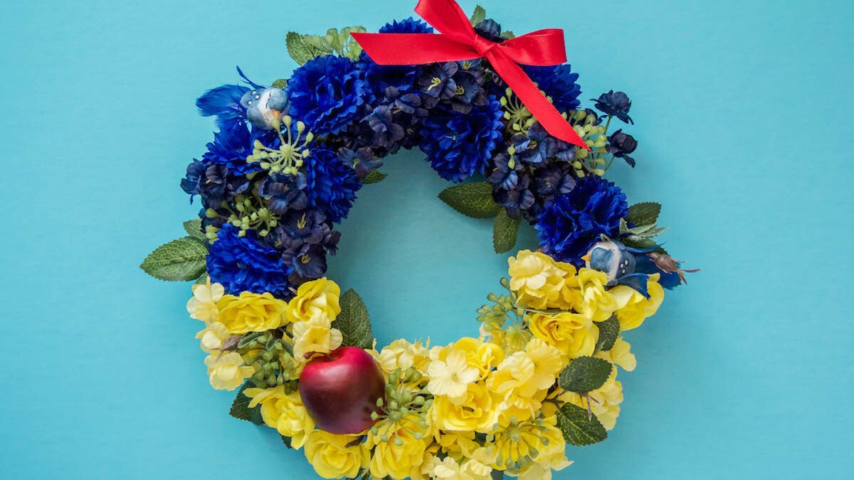 A Snow White themed wreath with blue and yellow flowers, a red bow, and a red apple.