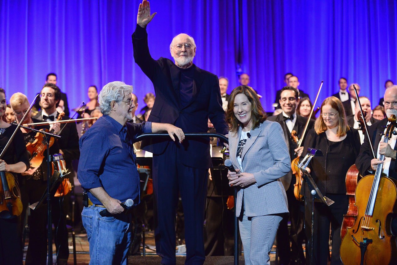 Standing on a podium between George Lucas and Kathleen Kennedy, John Williams waves to an unseen crowd while an orchestra stands behind him.