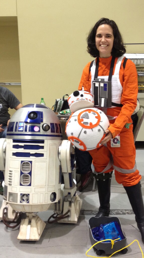 A fan in rebel pilot cosplay holding a crochet BB-8 next to a model of R2-D2.