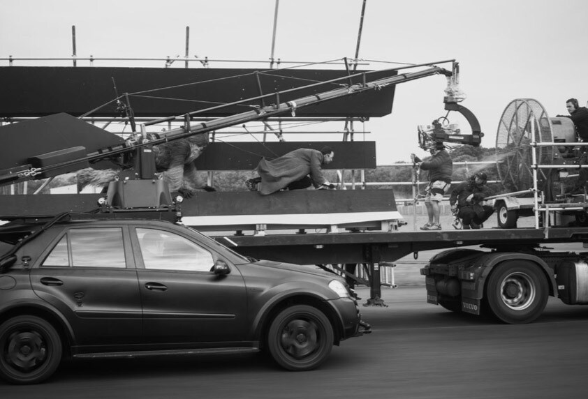 Stunt doubles crawl on the back of a large truck while a film crew sets up a shot in a behind-the-scenes image from the making of Solo: A Star Wars Story.