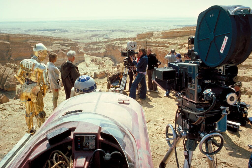 A film crew in the desert captures Luke, Obi-Wan, R2-D2, and C-3PO during filming of Star Wars: A New Hope.