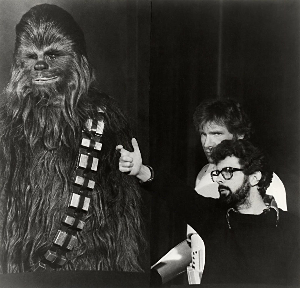 Peter Mayhew, in full costume as Chewbacca, taking direction from George Lucas alongside Harrison Ford