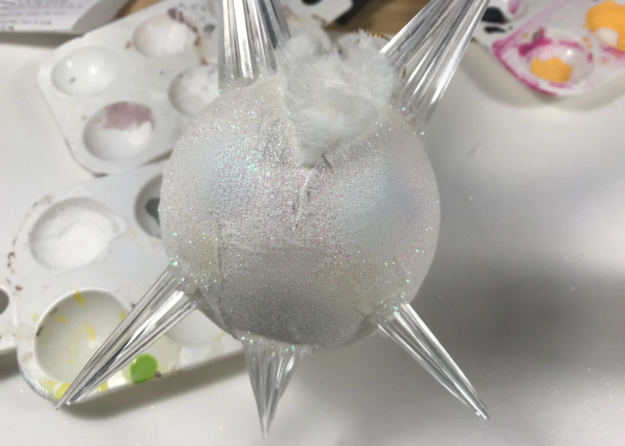 An unfinished homemade vulptex Christmas ornament. Glitter, craft fur, and clear toothpicks have have been added to a bulb ornament painted white.
