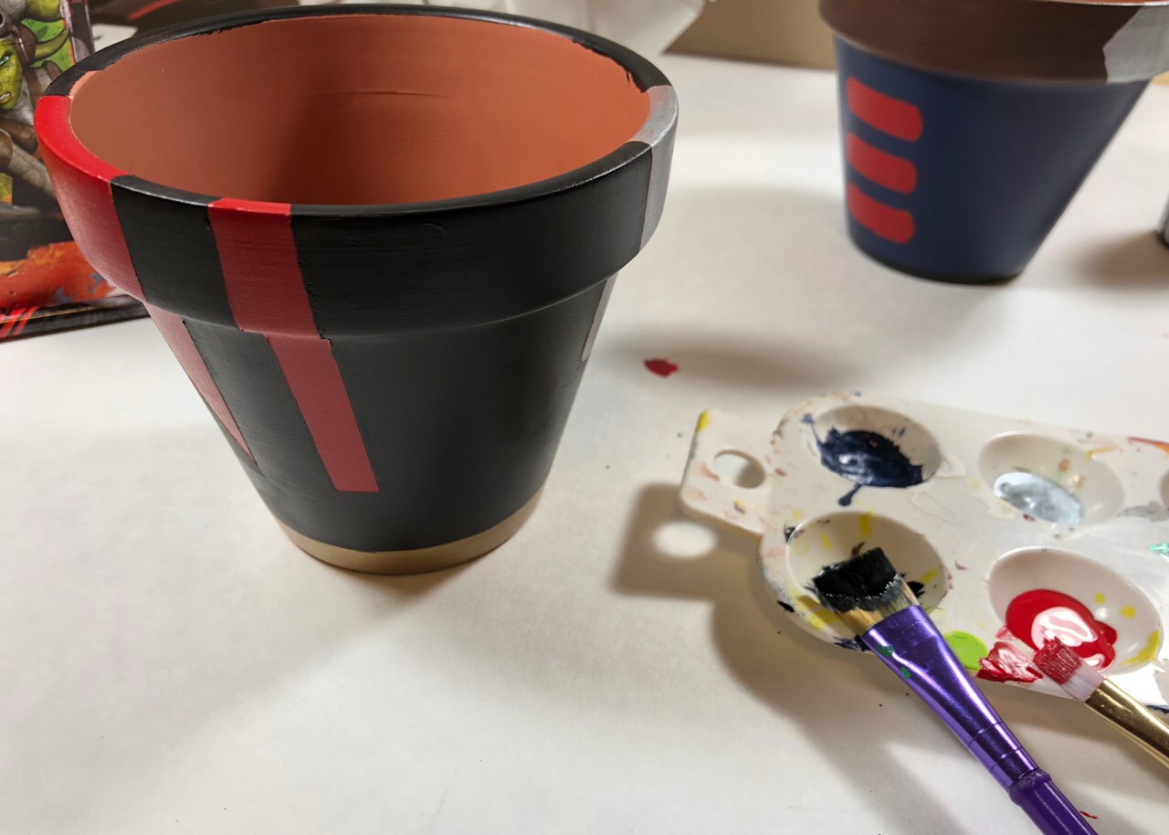 A terracotta pot painted black with red stripes to make it Star Wars themed, with a tray of paint and paint brushes next to it.