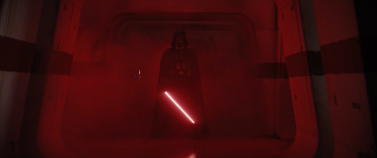 Darth Vader stands in the shadows, illuminated by the red glow from his lightsaber, in Rogue One.