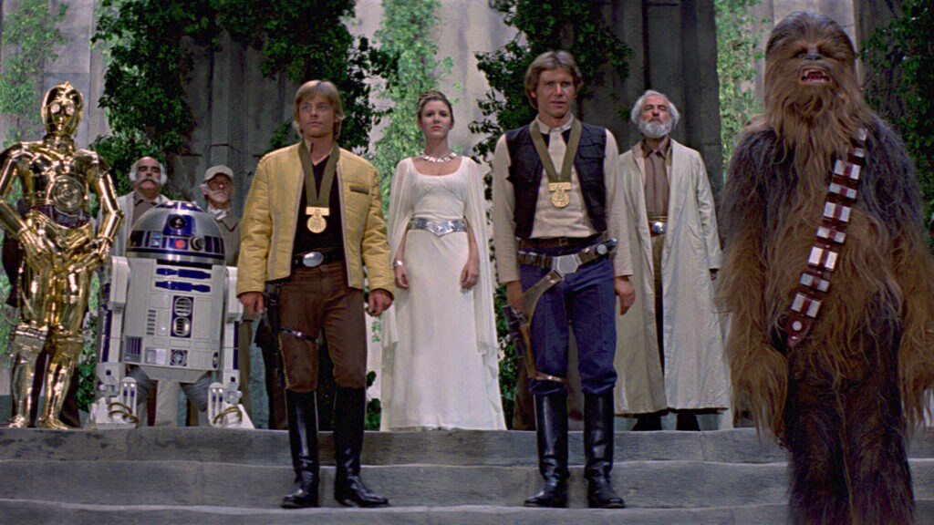 Luke and Han wear medals of Yavin as they stand with Leia, Chewie, C-3PO, and R2-D2 in the award ceremony after the defeat of the Death Star in A New Hope.
