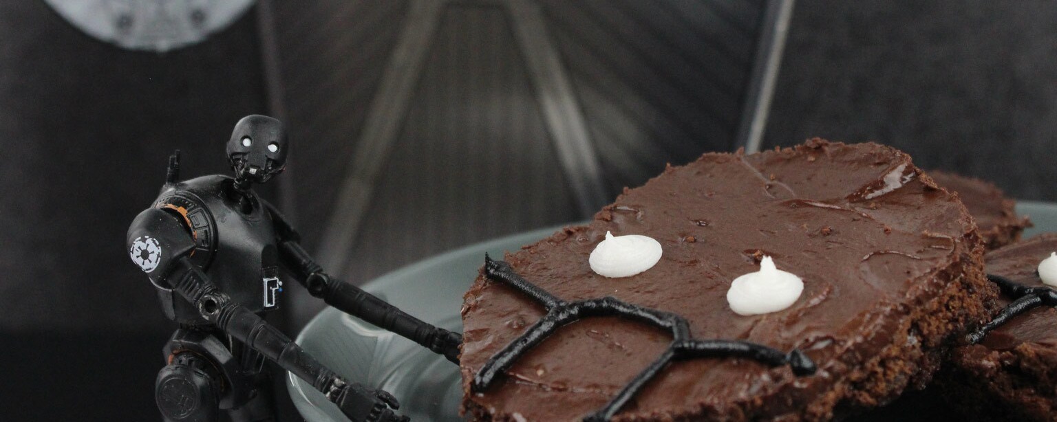 An action figure of security droid K-2SO stands next to a plate of moist fudge brownies which are decorated with frosting to replicate his face.