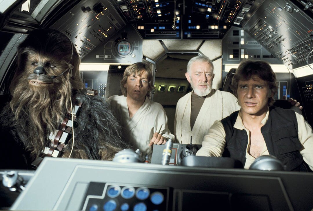 Han, Chewbacca, Luke, and Obi-Wan sit in the cockpit of the Millennium Falcon in A New Hope.
