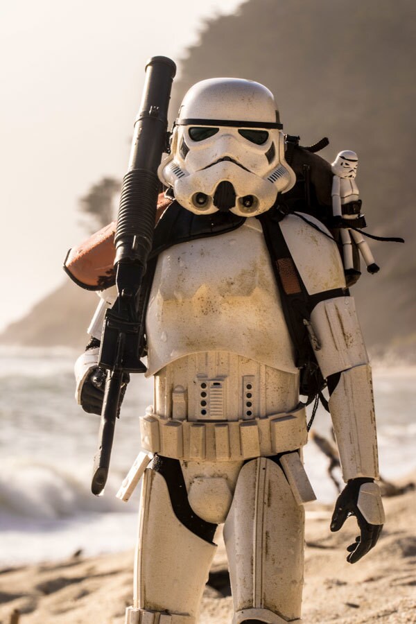 A stormtrooper action figure carries a blaster rifle and Jyn Erso's Stormie doll on a beach in a photograph by Johnny Wu.