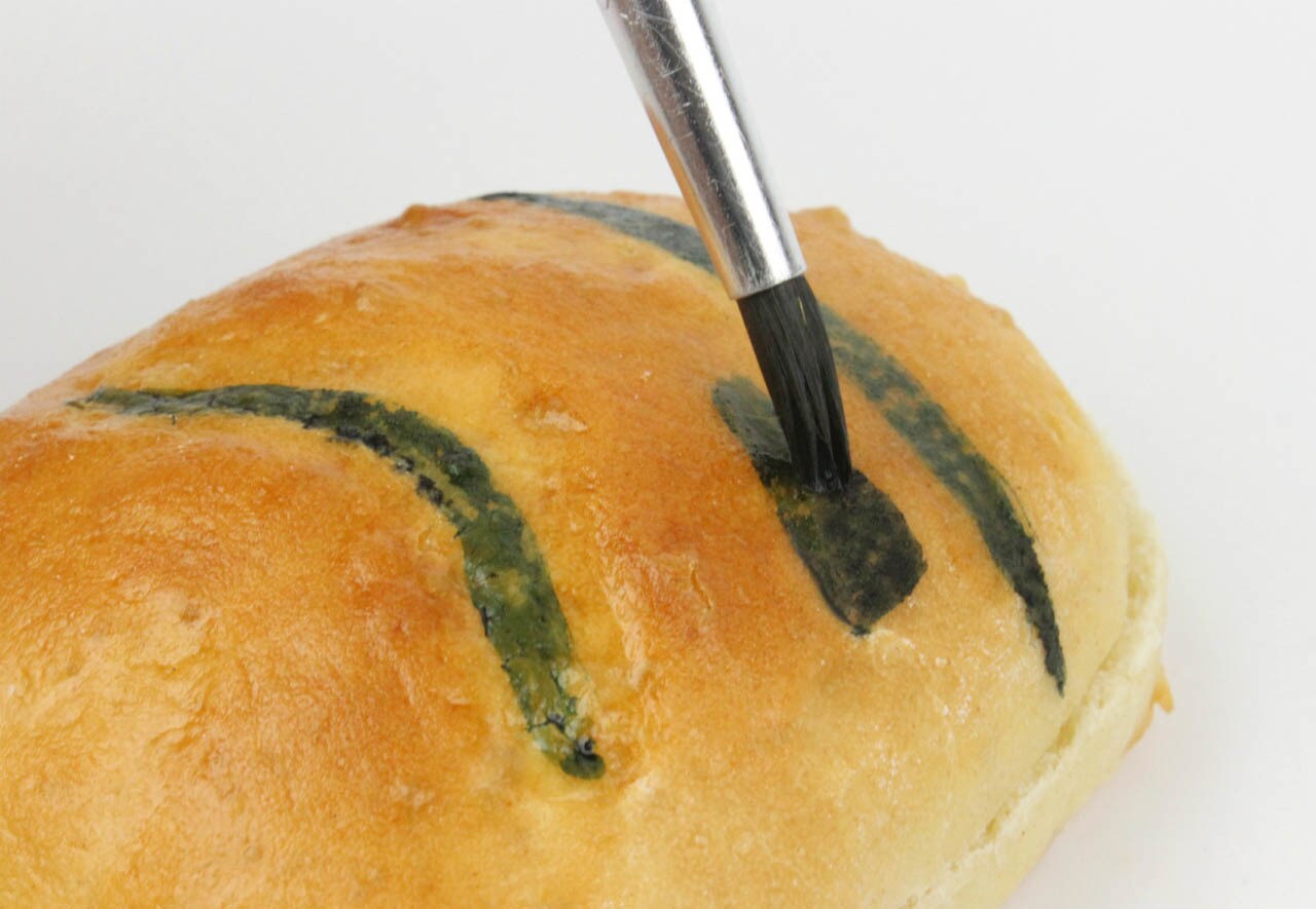 A kitchen brush paints black food gel dye onto a baked bun to create the look of a stormtrooper as part of the process for making Stormtrooper Sliders.