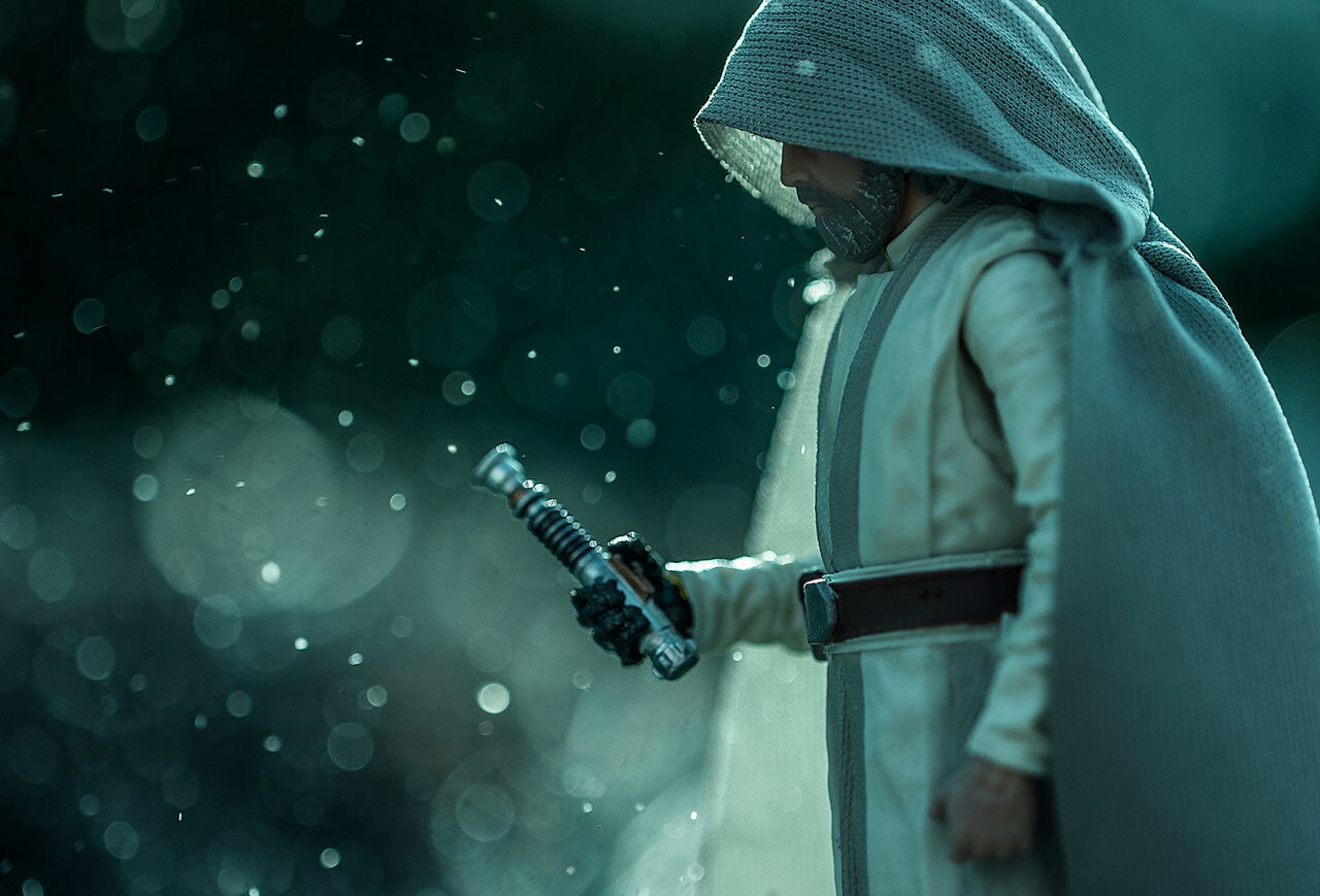 A cloaked and hooded action figure of Master Luke Skywalker holds a powered-down lightsaber in his gloved hand.