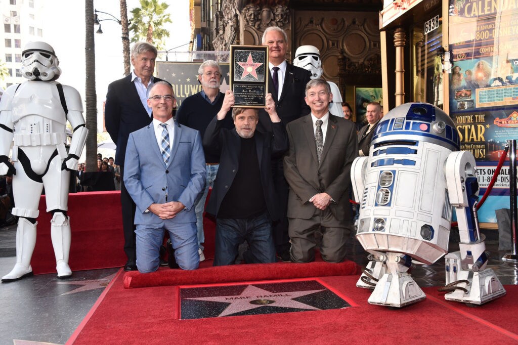 Mark Hamill shows the plaque he was presented with at a ceremony for the placement of his star on The Hollywood Walk of Fame. Also notably present are Harrison Ford, George Lucas, R2-D2, and a stormtrooper.