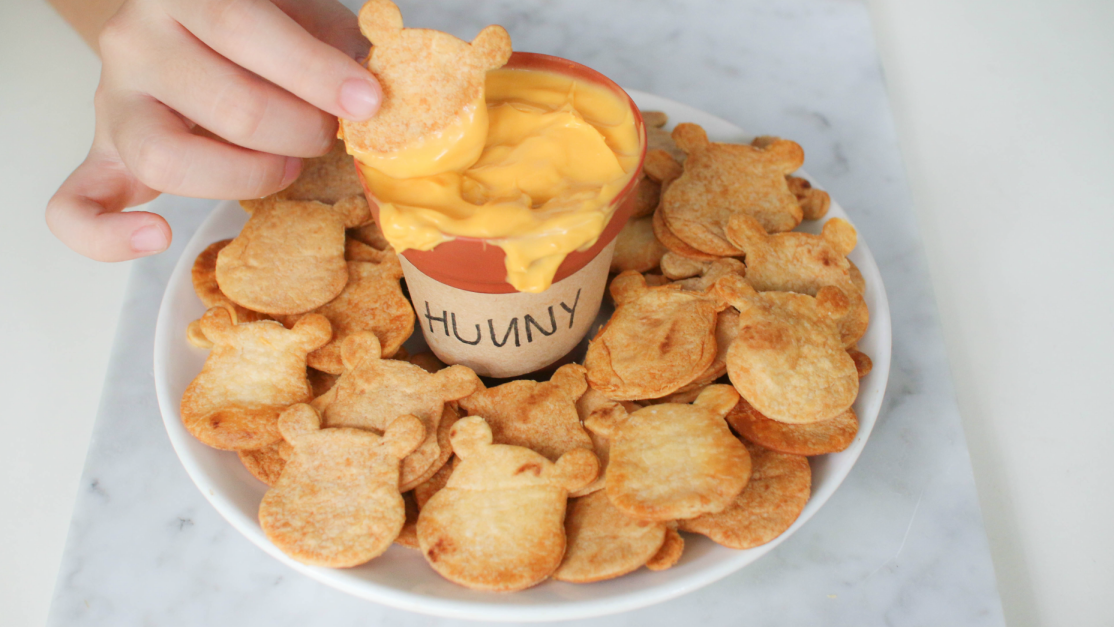 Winnie the Pooh shaped baked chips on a plate with cheese dip in a honey pot.