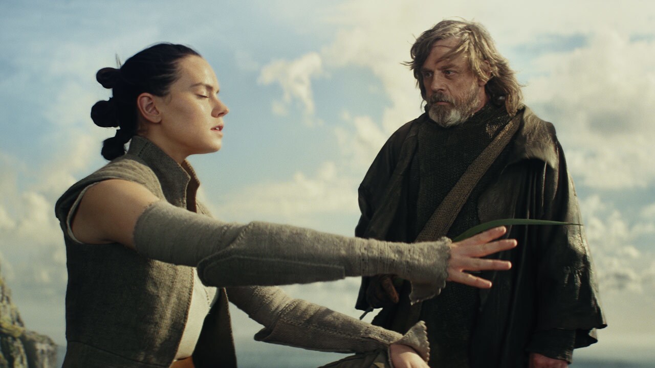 Rey closes her eyes with her arms extended in front of her as Luke watches in The Last Jedi.