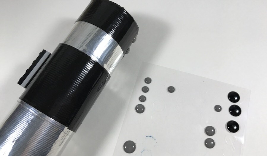 A vase partially decorated to resemble a lightsaber, next to a plastic sheet with black decorative dots attached.