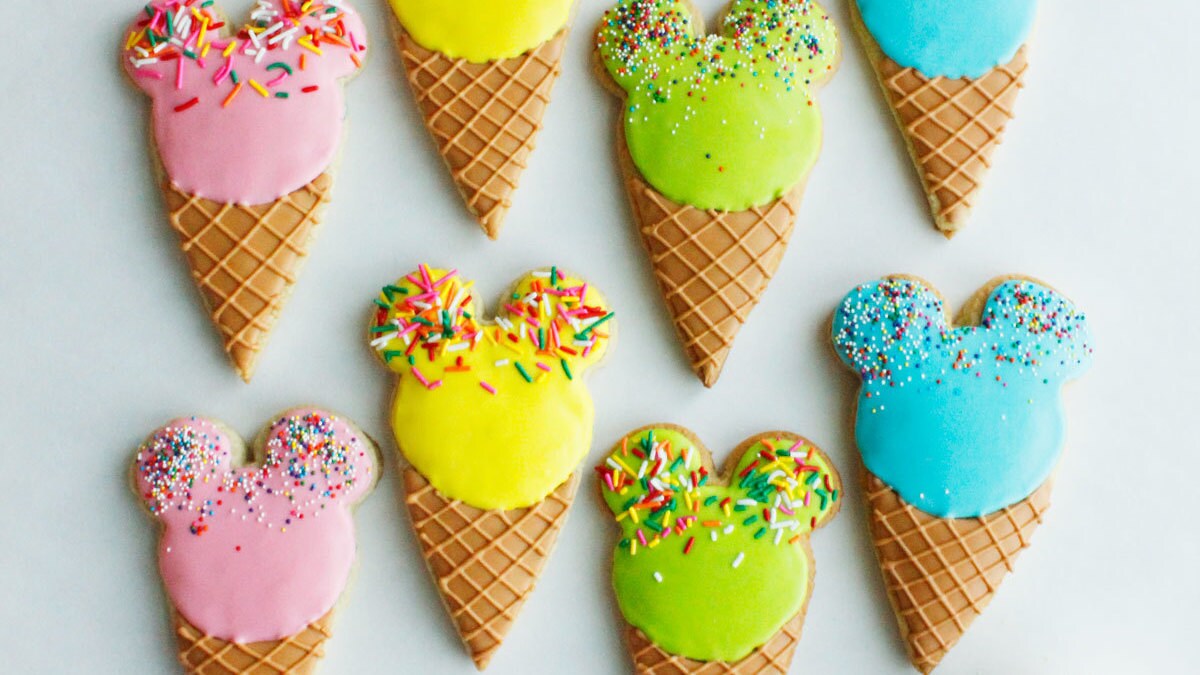 Several different colored cookies with Mickey Mouse frosting shaped like ice cream cones.