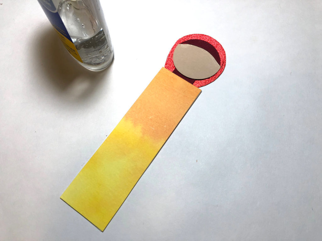 A round piece of tan paper outlined with red paper, which forms a handmaiden's head, is glued to the end of a rectangular piece of yellow and orange watercolor paper, which forms her body. This is a bookmark in progress.