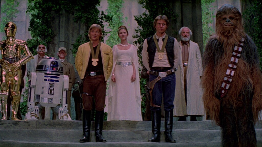 Luke and Han wear medals of Yavin as they stand with Leia, Chewie, C-3PO, and R2-D2 in the award ceremony after the defeat of the Death Star in A New Hope.