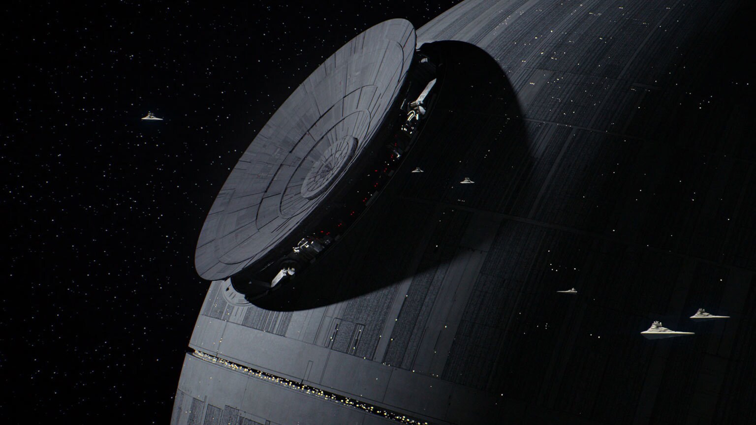 The Death Star as depicted in Rogue One, with several Star Destroyers appearing small in comparison as they pass in front of the station.