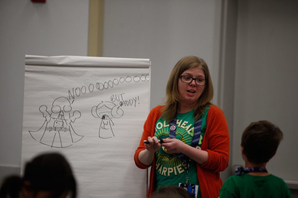A woman stands in front of an easel that has Darth Vader and Princess Leia drawn in black marker at a kid-friendly Star Wars Celebration class.