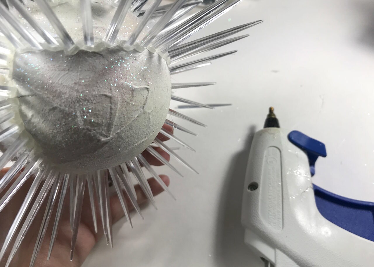 A hand holds an unfinished, homemade vulptex Christmas ornament next to a glue gun. Glitter, craft fur, and clear toothpicks have been added to a bulb ornament painted white.