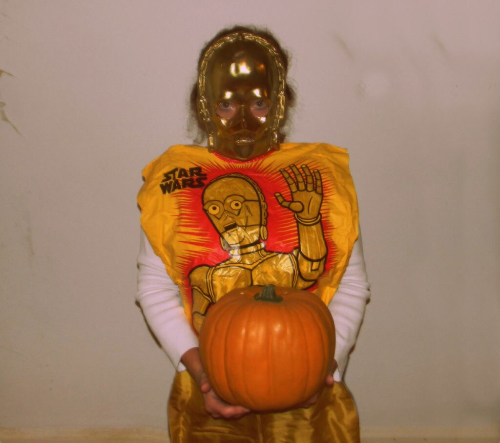 A child dressed in a C-3PO costume holds a pumpkin.