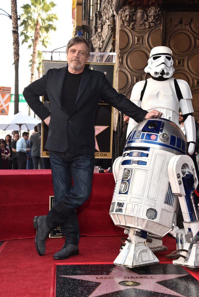 Mark Hamill poses for a photo with his hand on top of R2-D2's head while a stormtrooper stands behind at an event honoring him with a star on the Hollywood Walk of Fame.