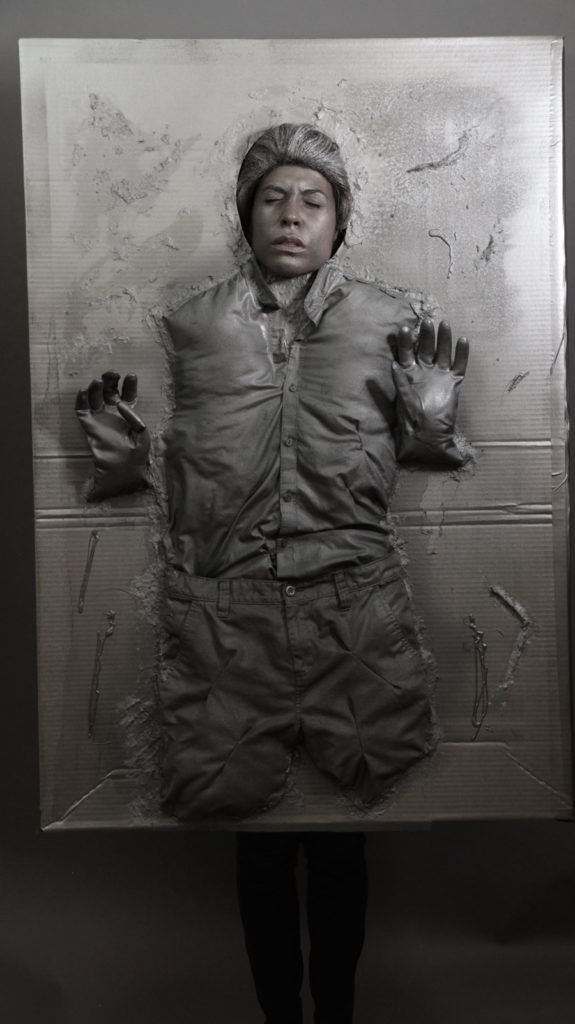 A person dressed as Han Solo in carbonite in a DIY Halloween costume made from a cardboard box, silver paint, an old shirt, and rubber gloves.