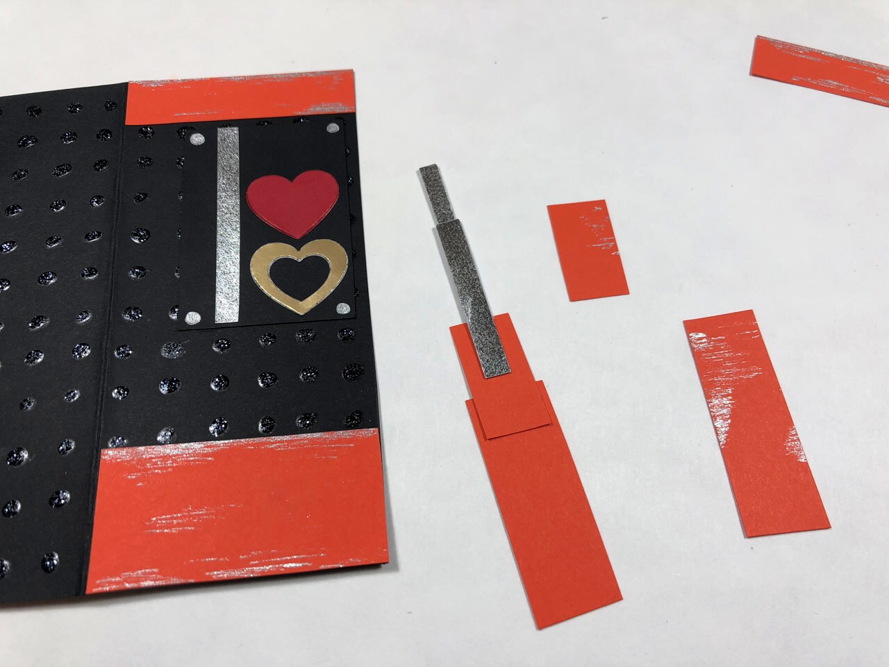 Black card stock with rows of black glitter glue, red and gold paper hearts, and strips of orange and silver cardstock beside additional strips of orange and silver cardstock. This is a Rose Tico-inspired Electro-Shock Prod Valentine's Day Card in progress.