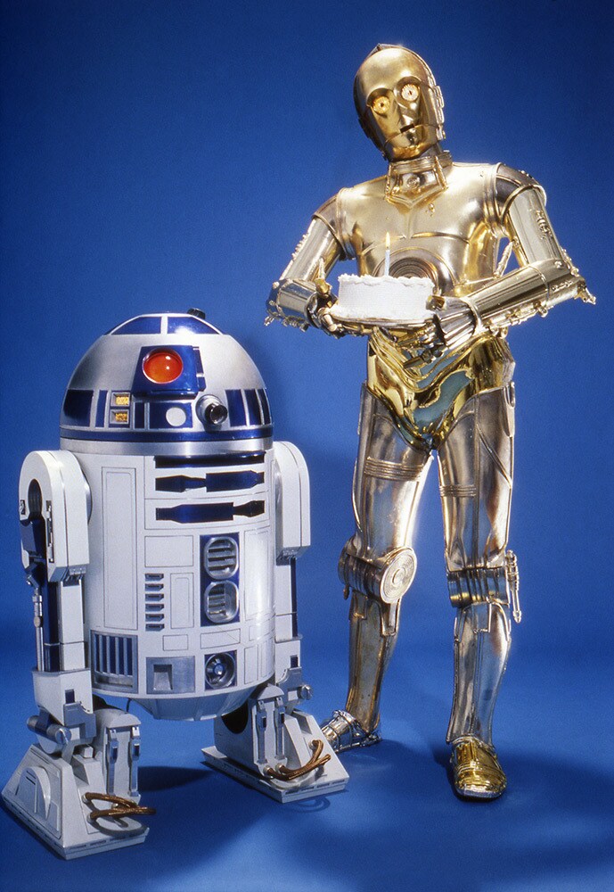 A photo of C-3PO, holding a cake with a lit candle, standing beside R2-D2.
