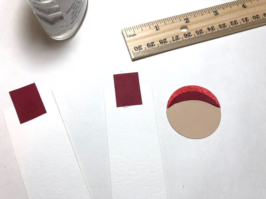 A round piece of tan paper with crescent-shaped pieces of red paper glued to one side, which forms a handmaiden's head, and two squares of red crepe paper glued to unpainted, rectangular strips of watercolor paper, which forms her body. This is a bookmark in progress.