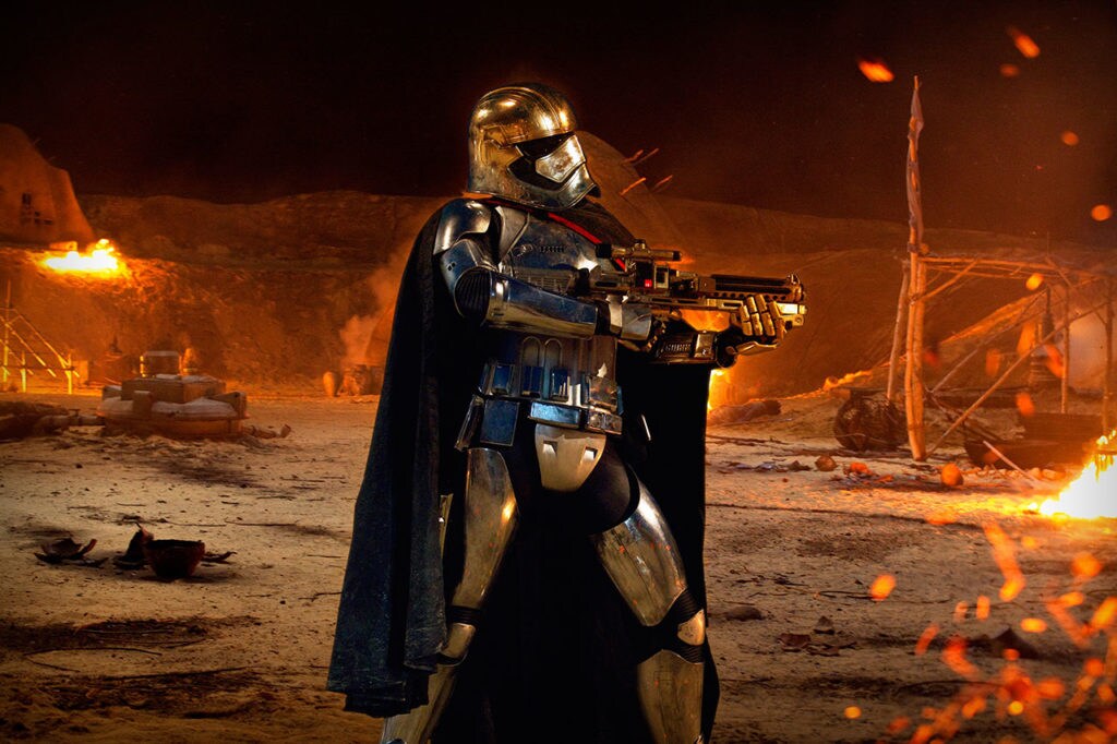 Captain Phasma wields her blaster rifle while standing in the middle of a burning village in Attack of the Clones.