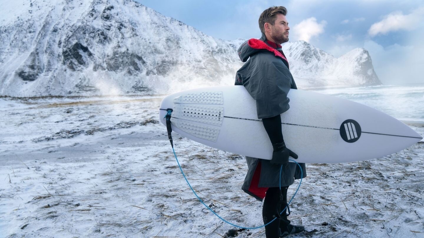 Chris Hemsworth stands with a surf board on a beach covered in snow