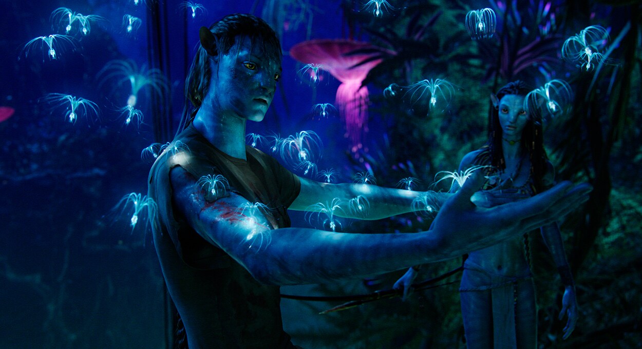 Jake Sully stands in the jungle on Pandora, with arms outstretched. Woodsprites land on his arms while Neytiri observes from the background.