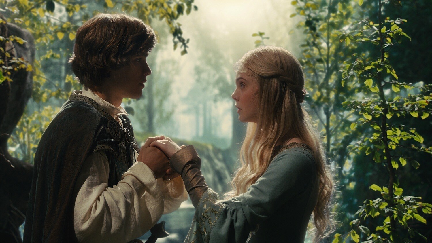 Elle Fanning as Aurora and Brenton Thwaites as Prince Phillip holding hands surrounded by trees in "Maleficent"