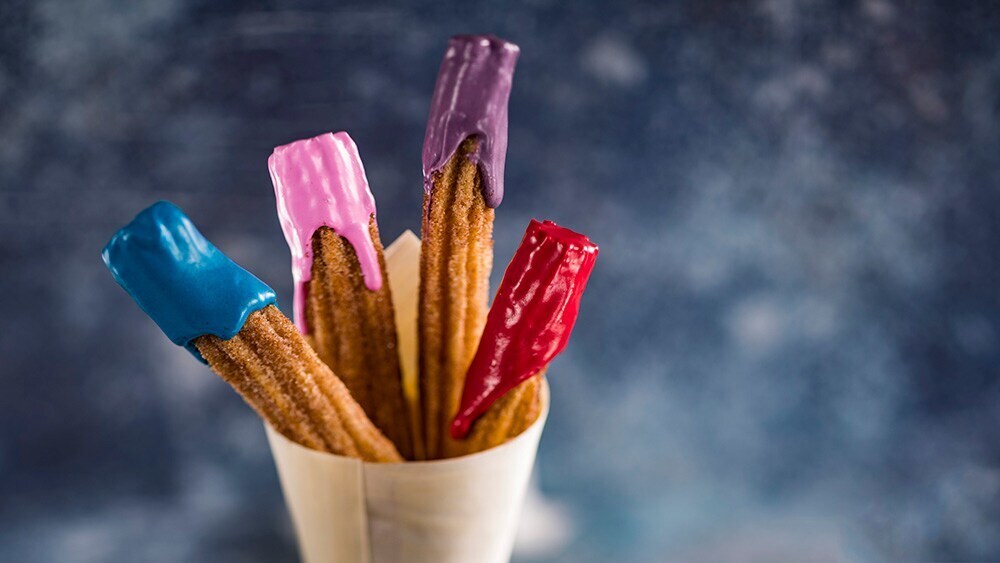 Four churros in a cone with dipped in different colors of frosting (l to r - blue, pink, purple, red)