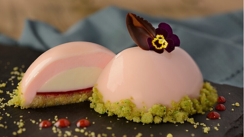 Image of a light pink vanilla, rose water and pistachio panna cotta that has a white layer, a thin red layer and a pistachio crumble along the rim