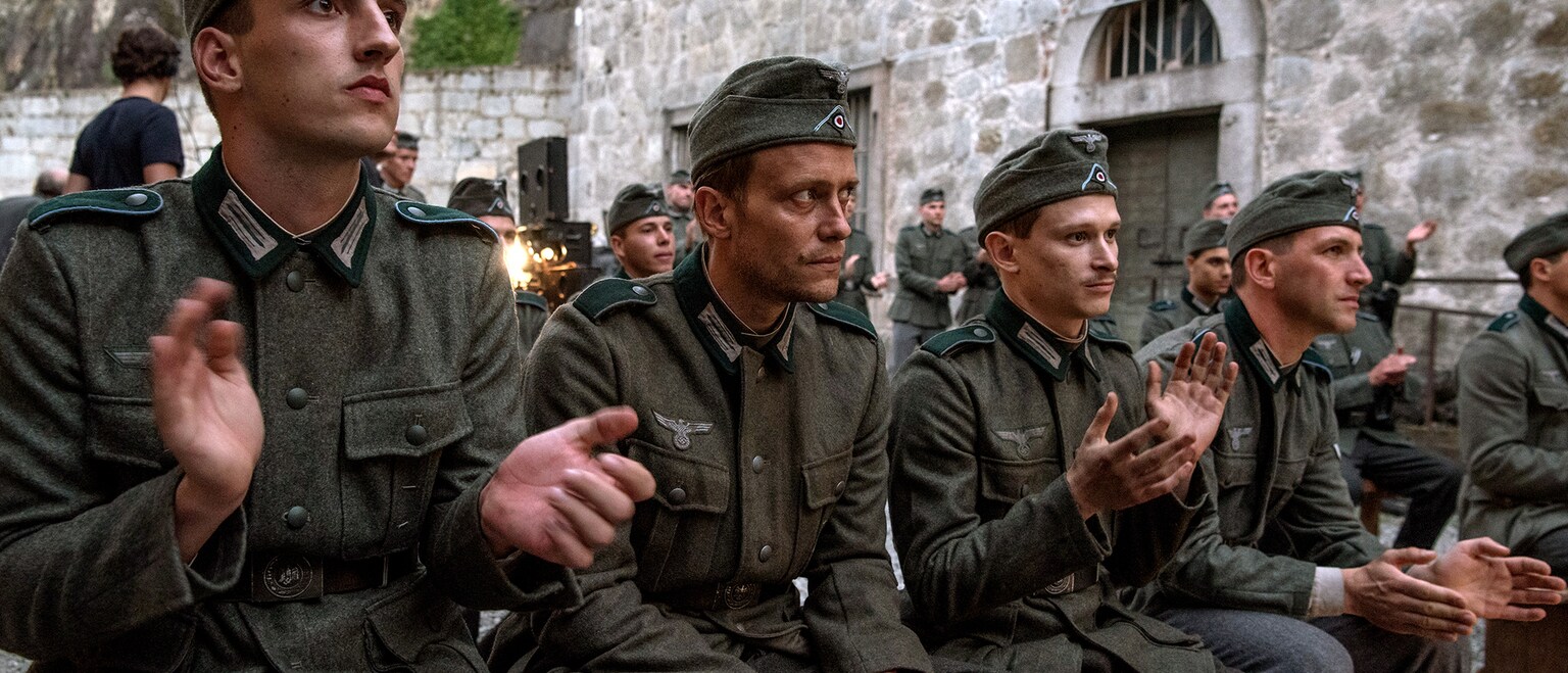 A German soldier sitting still while others clap to either side of him.
