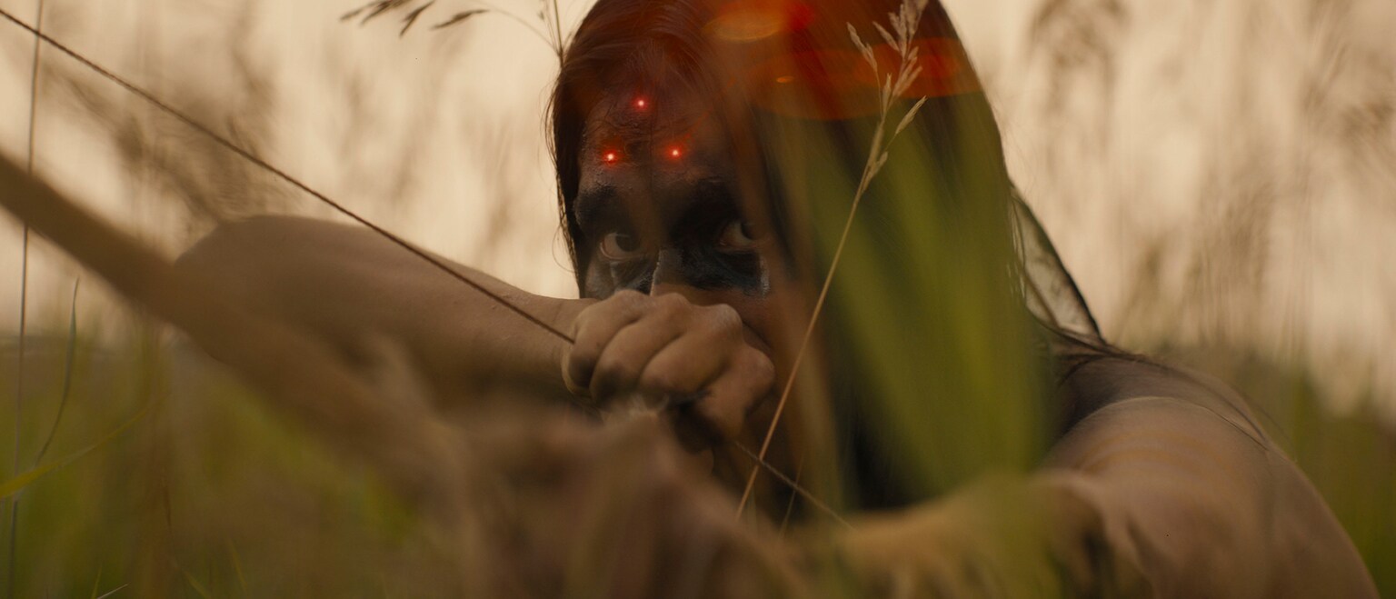 Image of a Native American with a bow drawn and a red laser sight on their forehead from the Hulu original movie, "Prey".