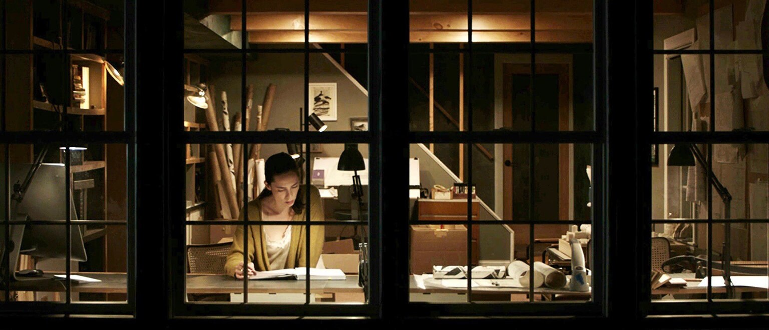 Image of a woman reading at a table through a window from the Searchlight's movie Night House.