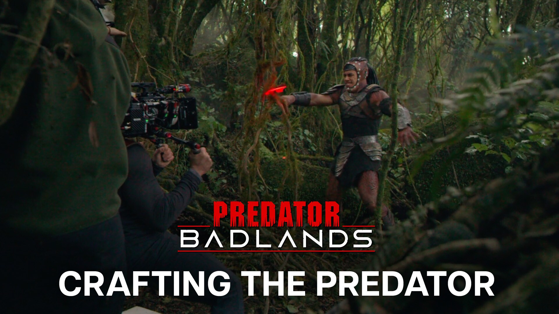 Image of an actor in a Predator costume being filmed by a camera crew in a woodland setting. The actor's face is visible as they are not wearing the full Predator mask. | Predator: Badlands | Crafting the Predator