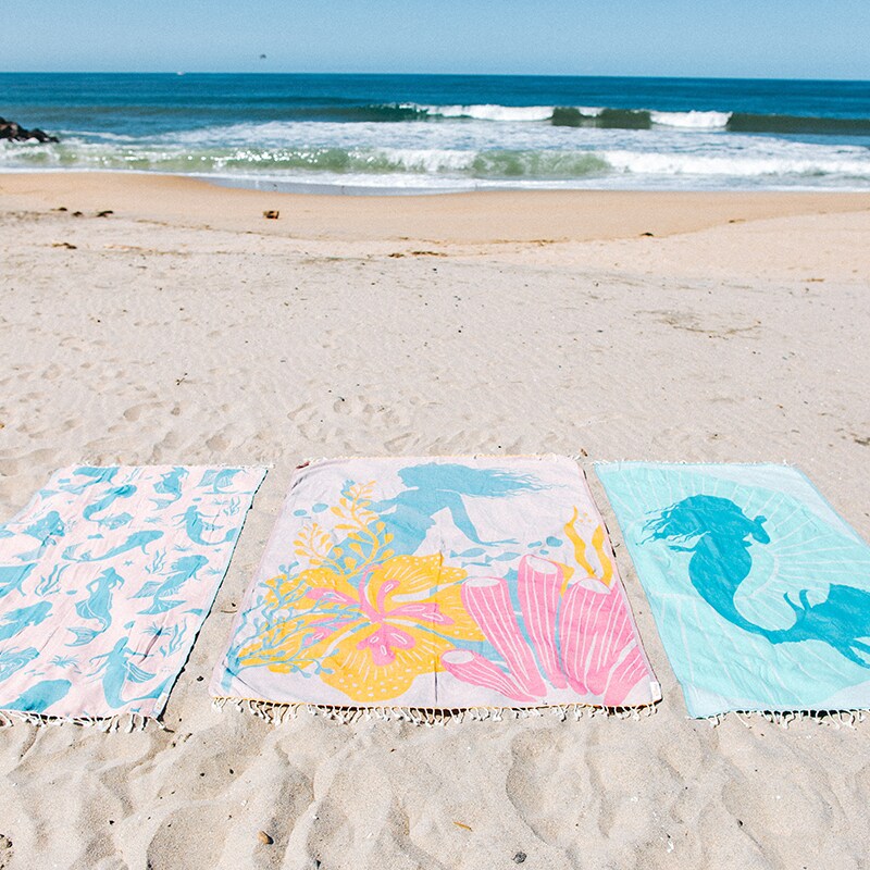 Image of three The Little Mermaid-inspired beach towels laid on the sand near the ocean.