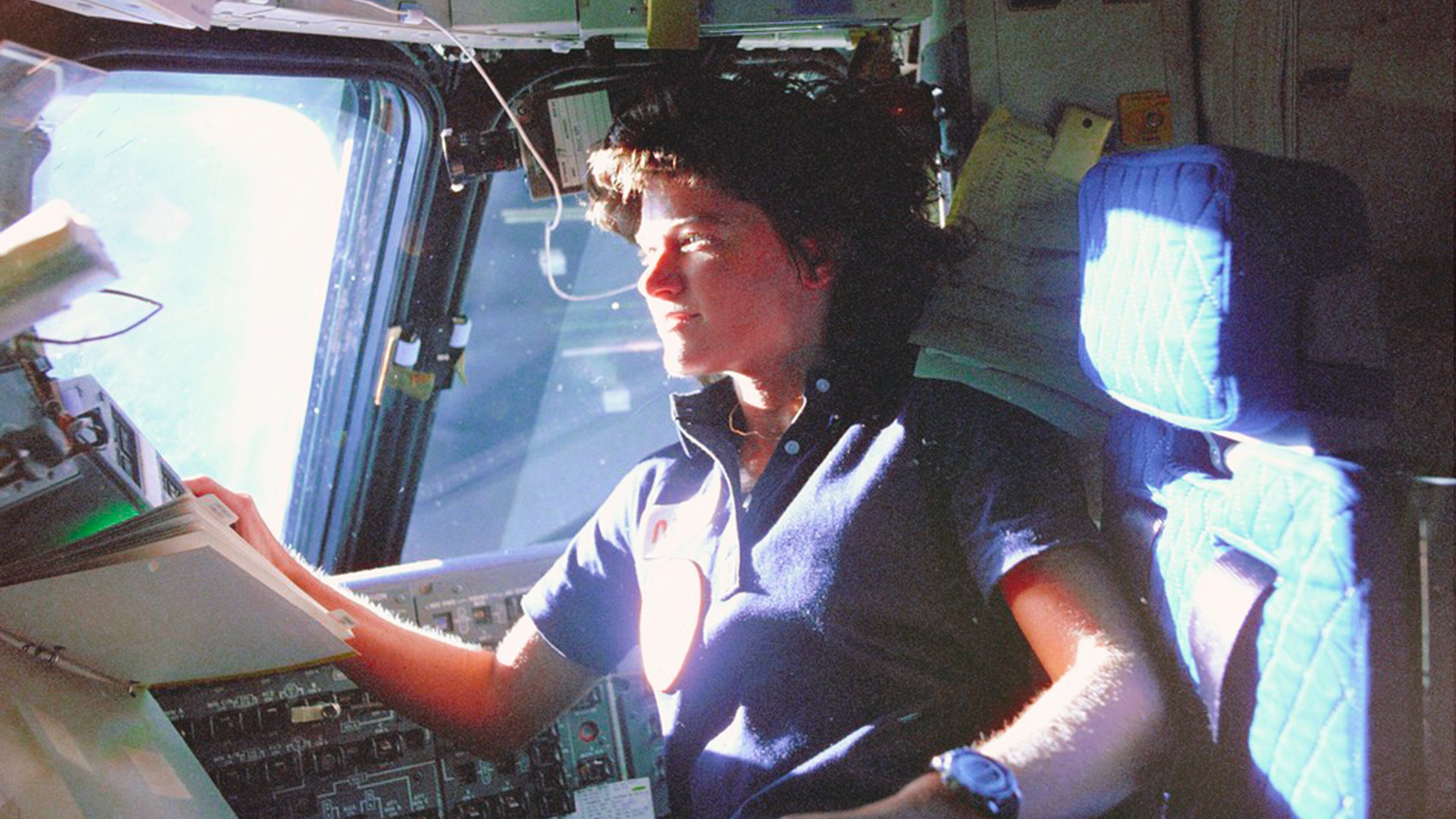 Astronaut Sally Ride, mission specialist on STS-7, monitors control panels from the pilot's chair on the Flight Deck. Floating in front of her is a flight procedures note book. (Credit: NASA)