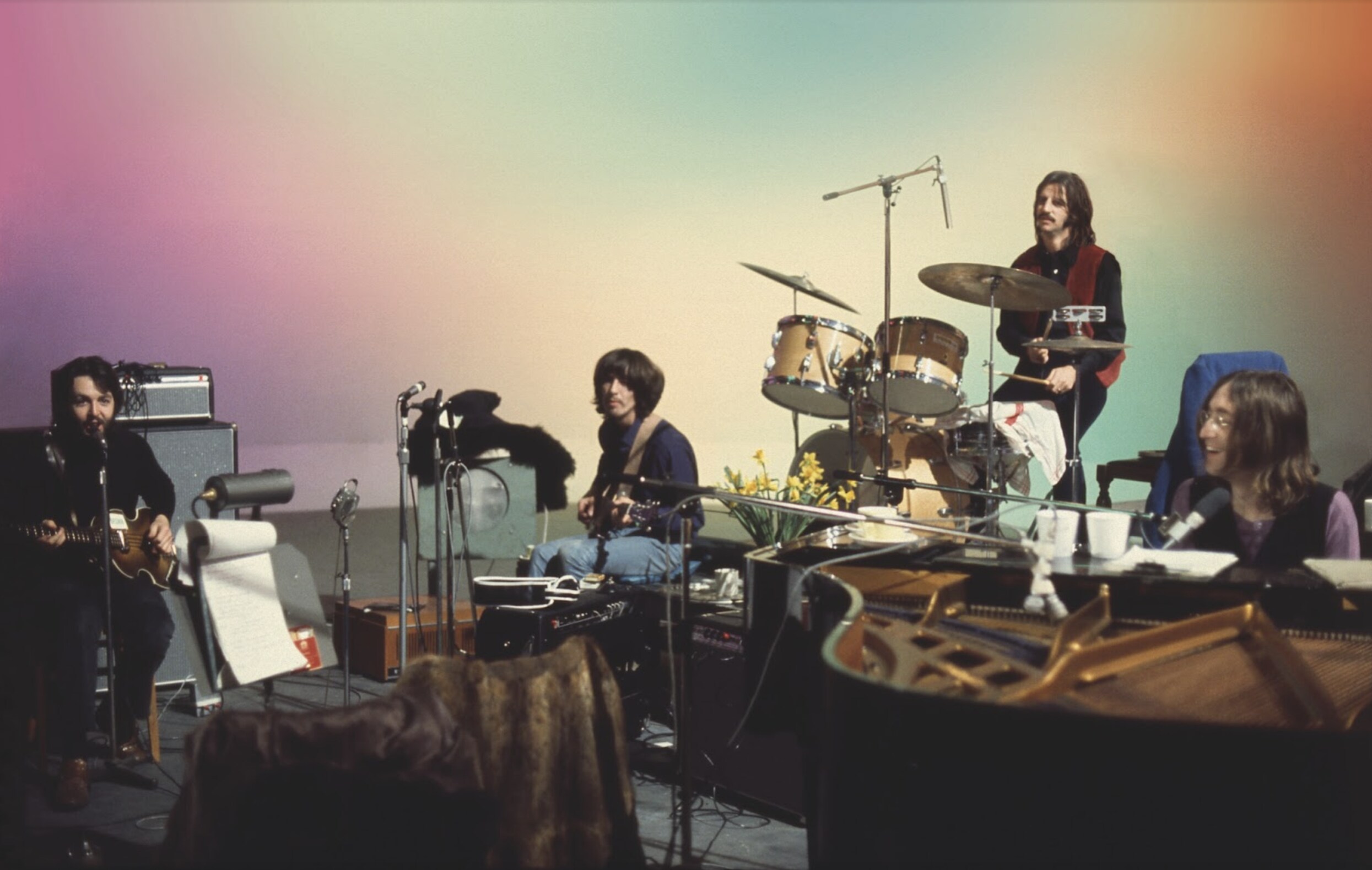 The Beatles perform on stage, each one playing a different instrument in front of a colored background 