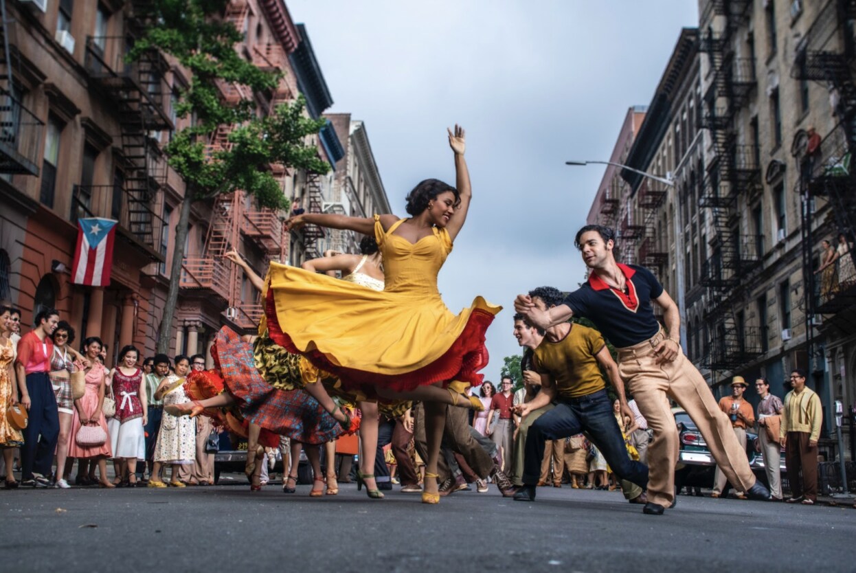 A girl in a yellow dress with a host of characters dance in the streets in West Side Story