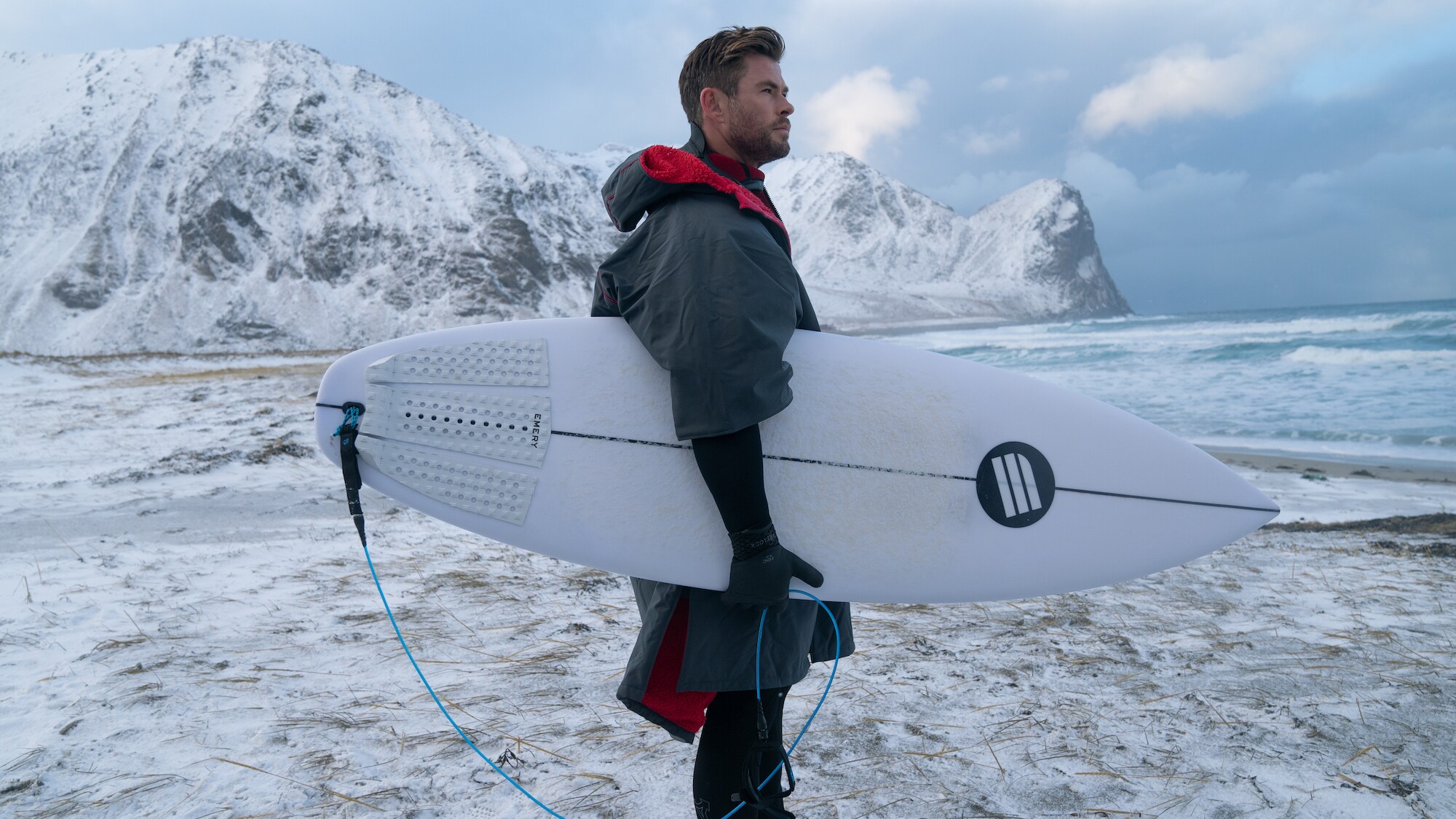 Chris Hemsworth stands on the beach with a surf board. (National Geographic for Disney+/Craig Parry)