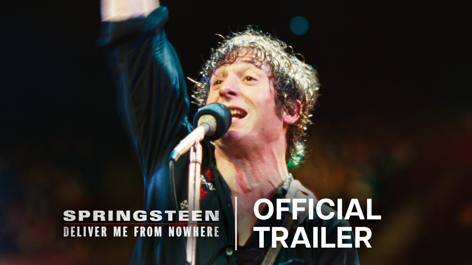 Image of Bruce Springsteen (actor Jeremy Allen White) on a stage with a raised right arm while standing in front of a microphone from the 20th Century Studios movie, "Springsteen: Deliver Me From Nowhere."