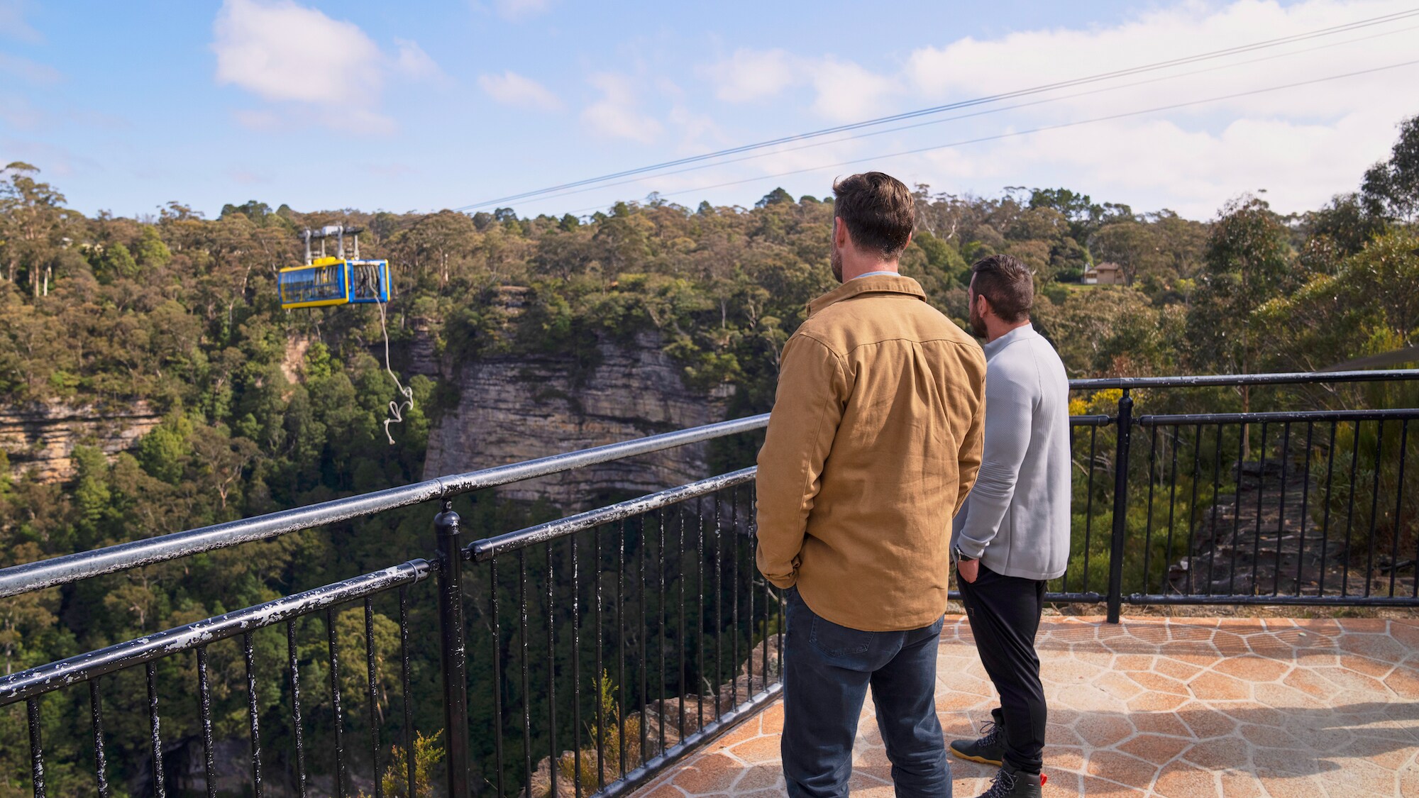Chris Hemsworth and sports scientist Ross Edgley gaze out at a cable cart suspended over a canyon with a 100-foot-long rope hanging from it. Chris will train to climb this rope with help from Ross.  (National Geographic for Disney+/Craig Parry)