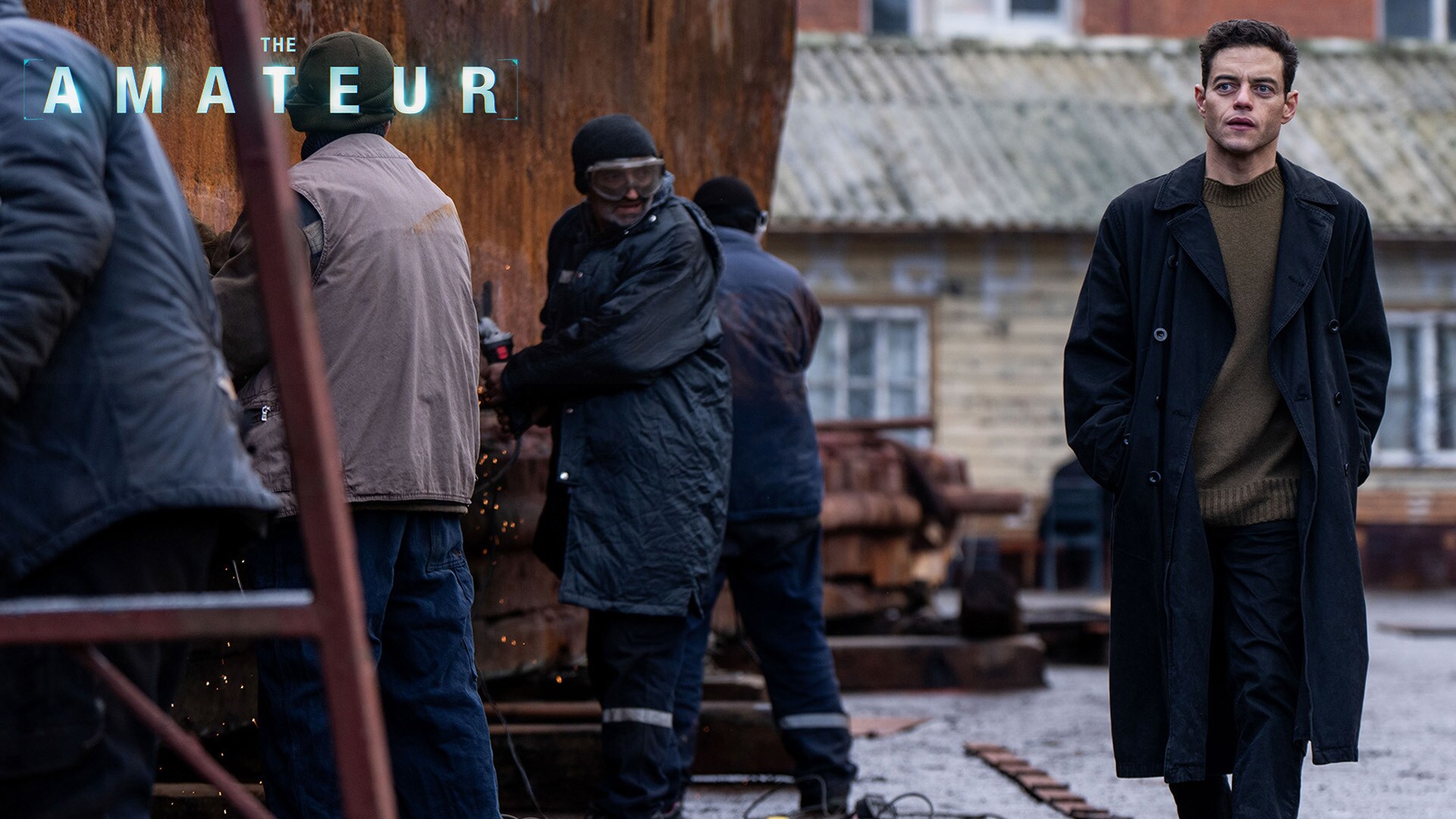 Image of actor Rami Malek wearing a trenchcoat walking by several individuals. | The Amateur